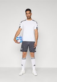 Bărbat îmbrăcat cu un tricou sport Adidas alb, pantaloni scurți gri și ghete albe, ținând o minge de fotbal Adidas albastră pe un fundal simplu.