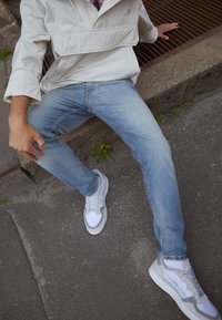 Světle modré džíny s úzkým střihem, doplněné bílými a šedými teniskami. Oblečení doplňuje béžová větrovka.