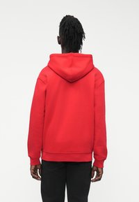 Κόκκινο βαμβακερό φούτερ με κουκούλα, σχεδιασμό τύπου πουλόβερ, ρυθμιζόμενο κορδόνι και τσέπη καγκουρό. Ριμπ μανσέτες και τελείωμα. Μαύρο παντελόνι φαίνεται από κάτω.