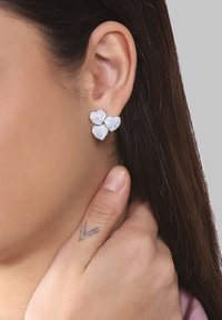 Silberne Ohrringe mit drei ineinandergreifenden Herzformen, von denen jede mit einem klaren Edelstein verziert ist, vor einem neutralen Hintergrund.