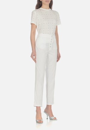 T-shirt bianco con piccoli dettagli metallici, abbinato a pantaloni bianchi a vita alta con chiusura a bottoni. Il modello indossa sandali con tacco argentati.