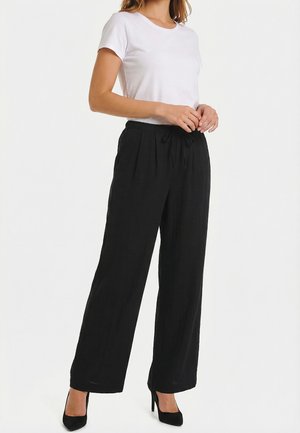 Femme portant un haut blanc à manches courtes, un pantalon ample noir taille haute avec un cordon de serrage, et des chaussures à talons noirs, debout devant un fond blanc.