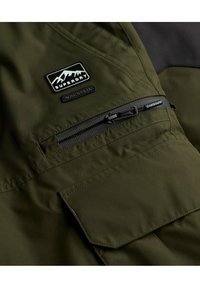 Donkergroene jas met een ritszak, gestructureerde stof en een zwart logo-patch met een bergontwerp. Glad oppervlak, waterafstotend materiaal.
