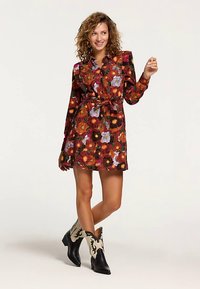 Vestito floreale con maniche lunghe, vita aderente, sfondo rosso intenso, fiori arancioni, gialli e viola. Paio di stivali da cowboy neri e crema.