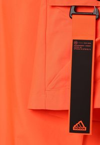 Oranžové sportovní šortky vyrobené z lehkého materiálu, s postranní kapsou a černým štítkem s logem Adidas a podrobnostmi o produktu.