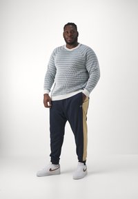 Suéter riscado azul e branco com textura canelada, combinado com calças de fato azuis-marinho com painel lateral bege. Usado com ténis brancos.