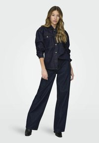 Camisa de mezclilla oscura tipo jacket con cierre de botones y dos bolsillos en el pecho, combinada con pantalones anchos de mezclilla oscura y botines negros.