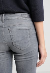 Harmaa farkkutakki, jossa on tiukka muoto. Ominaisuuksiin kuuluu takataskusuunnittelu ja vyötärönauhatunnus. Tekstuurissa on sileä pinta, jossa on hienovaraisia haalistumia.