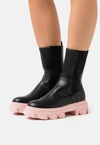 Botas de cuero negro hasta el tobillo con paneles elásticos y una suela gruesa rosa con una superficie texturada y un patrón de banda de rodadura definido.
