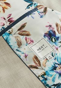Housse de vêtement florale avec un fond clair et des fleurs vibrantes bleues, roses et brunes. Comprend une étiquette blanche avec "NEXT TAILORING."