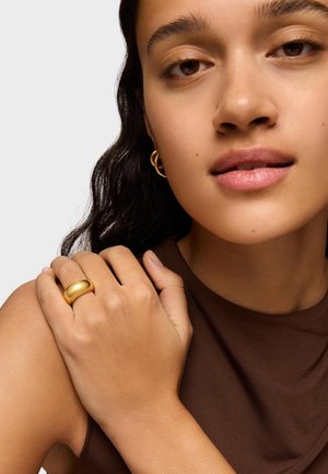 Gold-toned ring with a smooth, rounded design, worn on a hand resting on a shoulder; model has neutral makeup and dark, wavy hair.