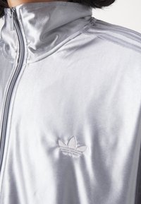Detailní záběr na lesklou stříbrnou bundu na zip s stojáčkovým límcem a vyraženým logem Adidas trefoil na hrudi.