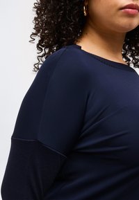 Γυναίκα με σγουρά μαλλιά που φοράει μπλε σκούρο μπλουζάκι με μακριά μανίκια και υφασμένο τελείωμα, εμφανίζεται από τον ώμο ως το πηγούνι πάνω σε απλό φόντο.