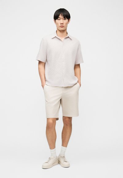 Άνδρας που φορά ανοιχτό μπεζ πουκάμισο με κοντά μανίκια και κουμπιά, μπεζ σορτς μέχρι το γόνατο, λευκές κάλτσες και μπεζ αθλητικά παπούτσια, στέκεται με τα χέρια στις τσέπες.