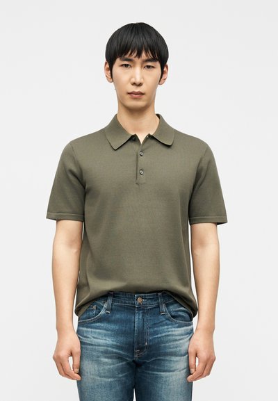 Jeune homme debout face à l'avant, portant un polo manches courtes vert olive et un jean bleu délavé sur un fond uni.