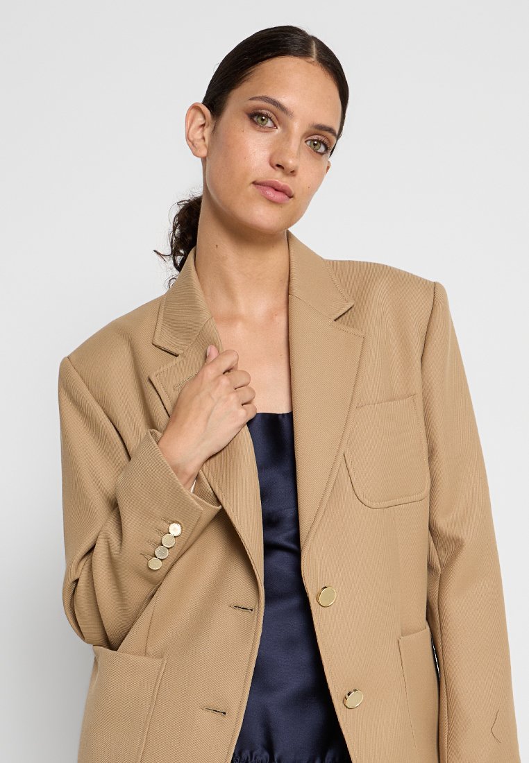 Beige blazer van getextureerde stof, grote notch-lapels, voorzakken en gouden knoopaccenten. Ggedragen over een donkere satijnen top.