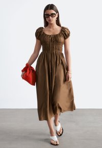 Femme portant une robe midi marron à manches courtes bouffantes, des sandales blanches, des lunettes de soleil foncées et tenant un sac rouge tout en marchant à l'intérieur.