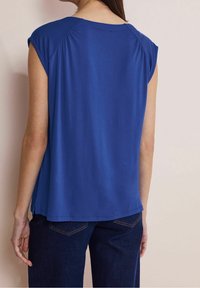 Royal blue short-sleeve top, smooth fabric, round neckline, relaxed fit, side slits at the hem, worn with dark blue jeans.
