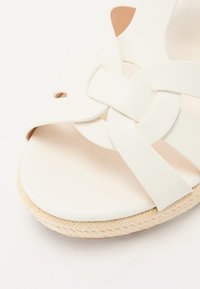 Vit sandal med flätade remmar, slät ovandel och en naturlig rep-sula för textur. Kurvad fotbädd för komfort.