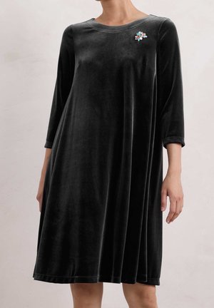Femme portant une robe en velours noire longueur genou avec des manches trois-quarts et une broche en pierres précieuses colorées sur le côté gauche de la poitrine.