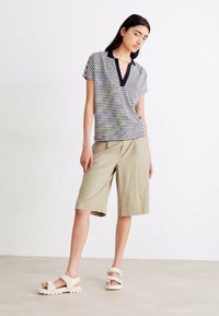 Stripete svart og hvit kortermet polo-skjorte, beige shorts og hvite sandaler, stående på en minimalisk bakgrunn.
