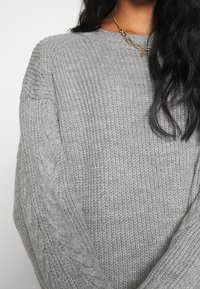 Femme portant un pull en maille gris clair texturé avec un motif torsadé sur les manches et un collier chaîne en or.