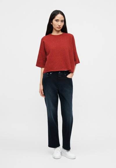 Κόκκινο κοντό πουλόβερ με κοντά μανίκια, σε συνδυασμό με σκούρο μπλε τζιν με ίσια πόδια και λευκά αθλητικά παπούτσια. Απλό φόντο, πλήρης προβολή σώματος.