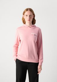 Sudadera rosa claro con un bolsillo delantero, capucha ajustable y un pequeño logo en el pecho. La tela parece suave y lisa.