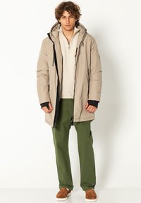 Beige gepolsterte Parka mit einem schwarzen Reißverschluss, verstellbarer Kapuze und gestrickten Bündchen, kombiniert mit einem hellen Pullover und grünen Hosen, dazu braune Schuhe.