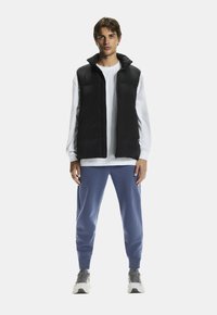 Czarna pikowana kamizelka na białej koszuli z długim rękawem, w połączeniu z niebieskimi dresami i szarymi trampkami, stojąca na tle białym.