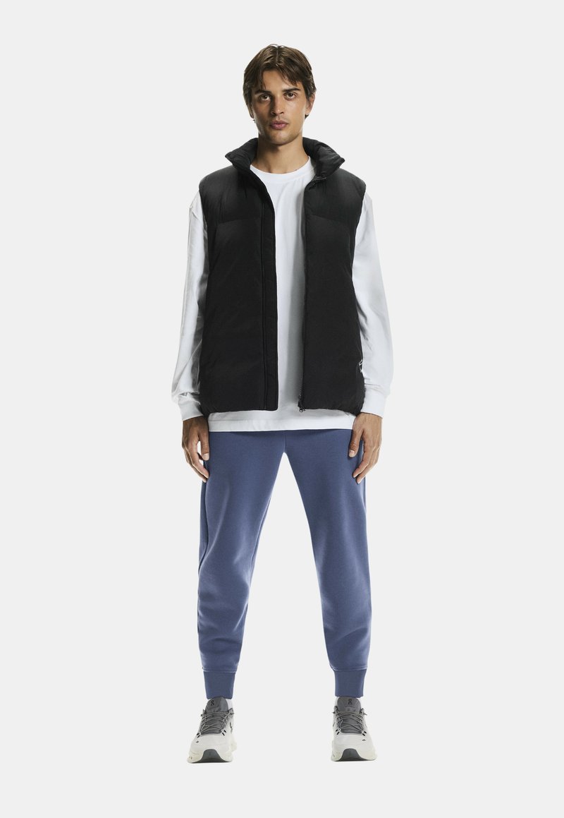 Czarna pikowana kamizelka na białej koszuli z długim rękawem, w połączeniu z niebieskimi dresami i szarymi trampkami, stojąca na tle białym.