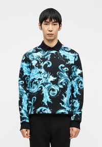 Άνδρας που φοράει μαύρο μακρυμάνικο πόλο με φωτεινό μπλε παλλόμενο μοτίβο νερού, στέκεται μπροστά σε απλό λευκό φόντο, κοιτάζοντας προς τα εμπρός.