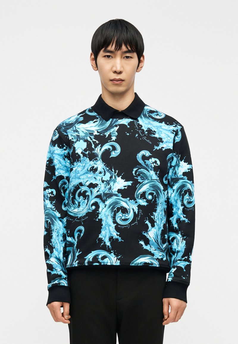 Άνδρας που φοράει μαύρο μακρυμάνικο πόλο με φωτεινό μπλε παλλόμ ενο μοτίβο νερού, στέκεται μπροστά σε απλό λευκό φόντο, κοιτάζοντας προς τα εμπρός.