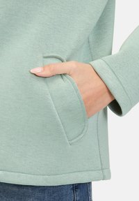 Jasnozielony sweter z gładką teksturą, z boczną kieszenią i zaokrąglonym dołem. Rękaw ma prosty, czysty wykończenie.