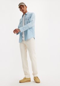 Lichtblauwe denim shirt met drukknopen, gecombineerd met crèmekleurige broek en tan suède schoenen. Een pet met logo maakt de casual outfit compleet.