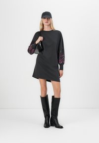 Svart sweatshirtkjole med blomstermønster på lange ermer, kombinert med høye svarte støvler og en svart caps. Liten håndveske i hånden.