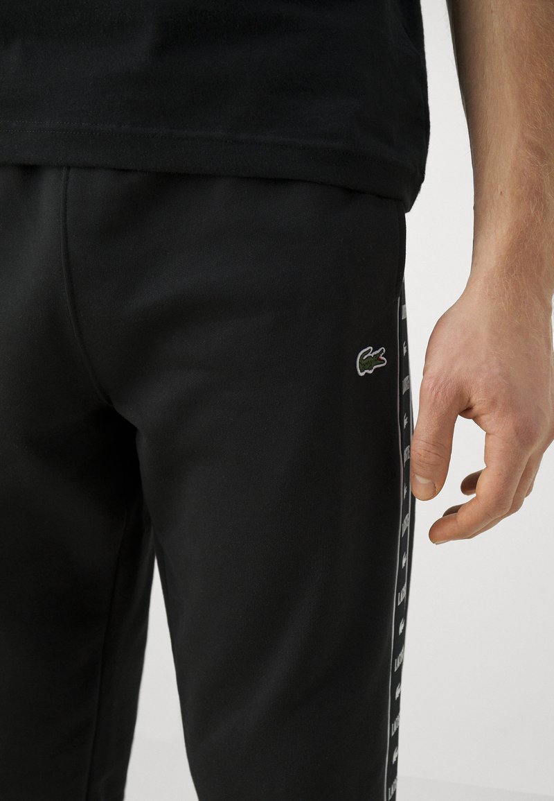 Czarne dresy o gładkiej fakturze, z małym zielonym logo krokodyla i białymi bocznymi paskami z powtarzającym się wzorem tekstu.