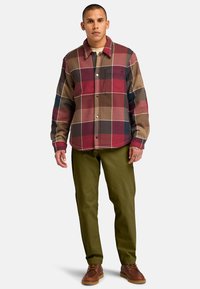 Camicia di flanella in un motivo a quadri marrone, rosso e blu marino, con chiusura a bottoni e taschino sul petto; abbinata a pantaloni verde oliva e scarpe marroni.