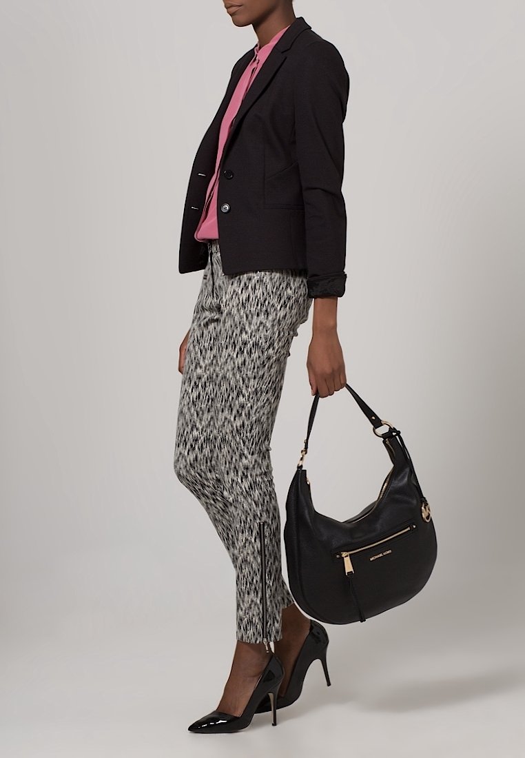 Femme en blazer noir, blouse rose, pantalon slim à motif, escarpins noirs, tenant un sac à main en cuir noir avec des détails dorés.