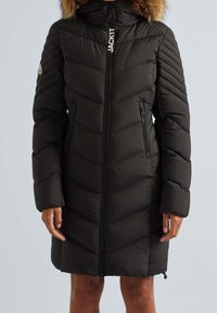 Schwarzer Steppmantel mit Chevron-Muster, ausgestattet mit einer Kapuze mit Fellbesatz, Reißverschlüssen vorne und an den Seiten sowie gerippten Bündchen.