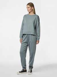 Kvinne iført en ledig, blågrønn genser og matchende joggebukser med svarte og hvite joggesko, stående mot en ensfarget bakgrunn.