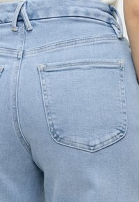 Ανοιχτό μπλε τζιν παντελόνι από την πίσω όψη που δείχνει ραφές, μία πίσω τσέπη και θηλιές ζώνης σε ένα άτομο που φοράει λευκό τοπ.