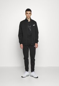 Black zip-up jacket and matching joggers made of soft fabric, featuring a PUMA logo, paired with white athletic shoes and a black t-shirt.