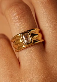 Goldfarbener Ring mit einer polierten Oberfläche, der eine rechteckige Edelsteinmitte, glatte abgerundete Kanten und geriffelte Akzente am Ringband aufweist.