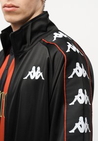 Νεαρό άτομο που φοράει μαύρο μπουφάν με φερμουάρ, στο οποίο υπάρχουν λευκά λογότυπα με καθρεφτισμένες φιγούρες καθιστών κατά μήκος του ώμου και του στήθους, πάνω από κόκκινη και μαύρη μπλούζα.