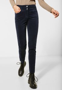 Marineblauwe corduroy broeken met slim fit, twee zijzakken, dubbele knoopsluiting en gestructureerde stof. G gedragen met zwarte veterschoenen.