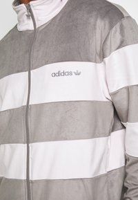 Grå och vit randig zip-up jacka tillverkad av mjukt tyg, med hög krage och broderad logotyp på bröstet.