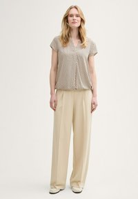 Blouse à manches courtes, à motifs, en beige avec un décolleté en V, associée à un pantalon large beige. Le modèle se tient devant un fond neutre.