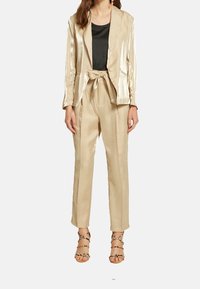 Ensemble blazer et pantalon en métal beige avec une taille nouée. Le blazer présente un col à revers, tandis que le pantalon a une coupe décontractée.