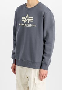 Sweat-shirt gris en tissu doux avec encolure ronde, poignets côtelés et un grand logo crème "ALPHA INDUSTRIES" sur le devant.
